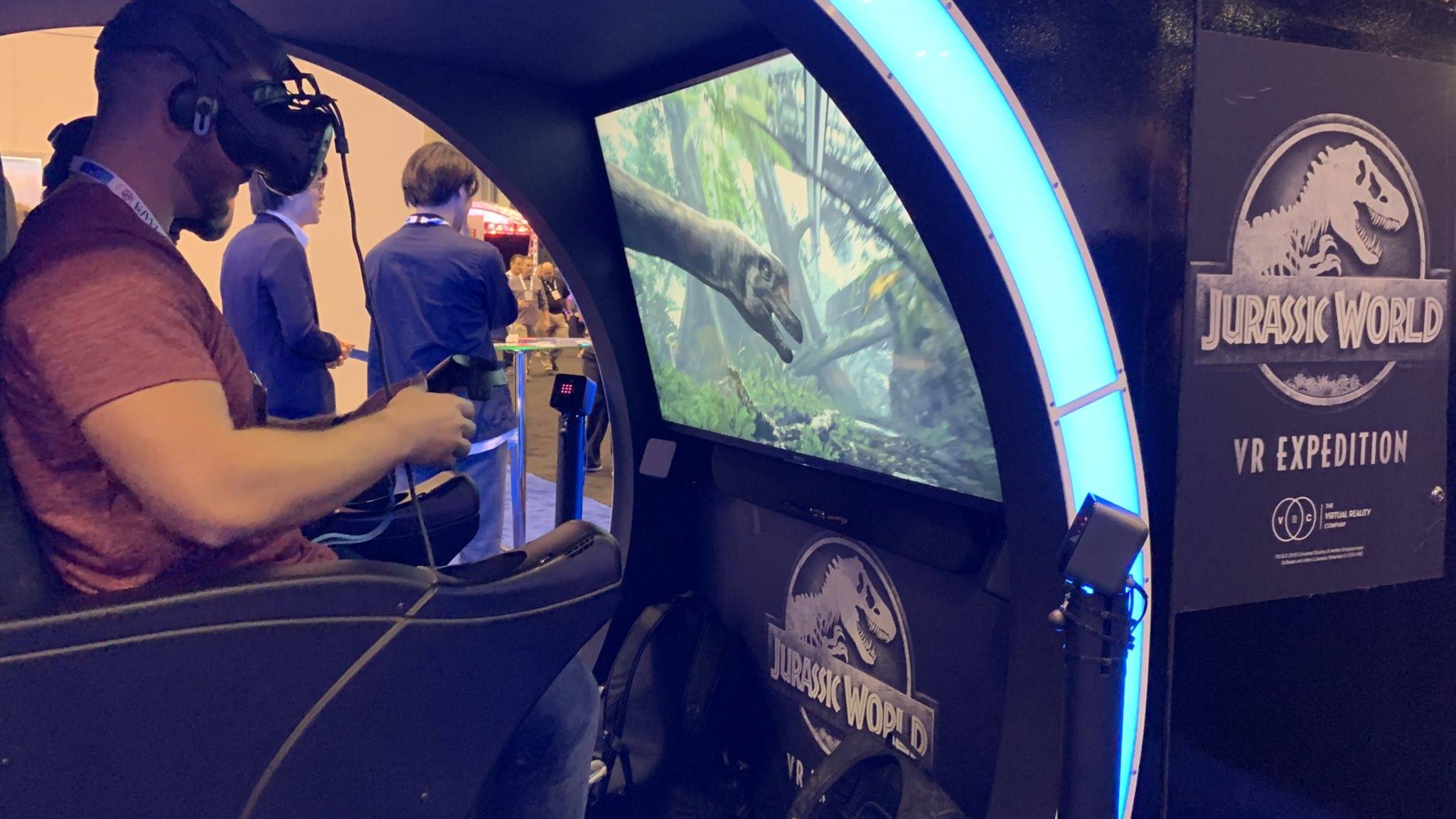 A rider in a VR headset in the Jurassic World VR Expedition motion-platform seat, with a dinosaur on the screen beside him, at IAAPA