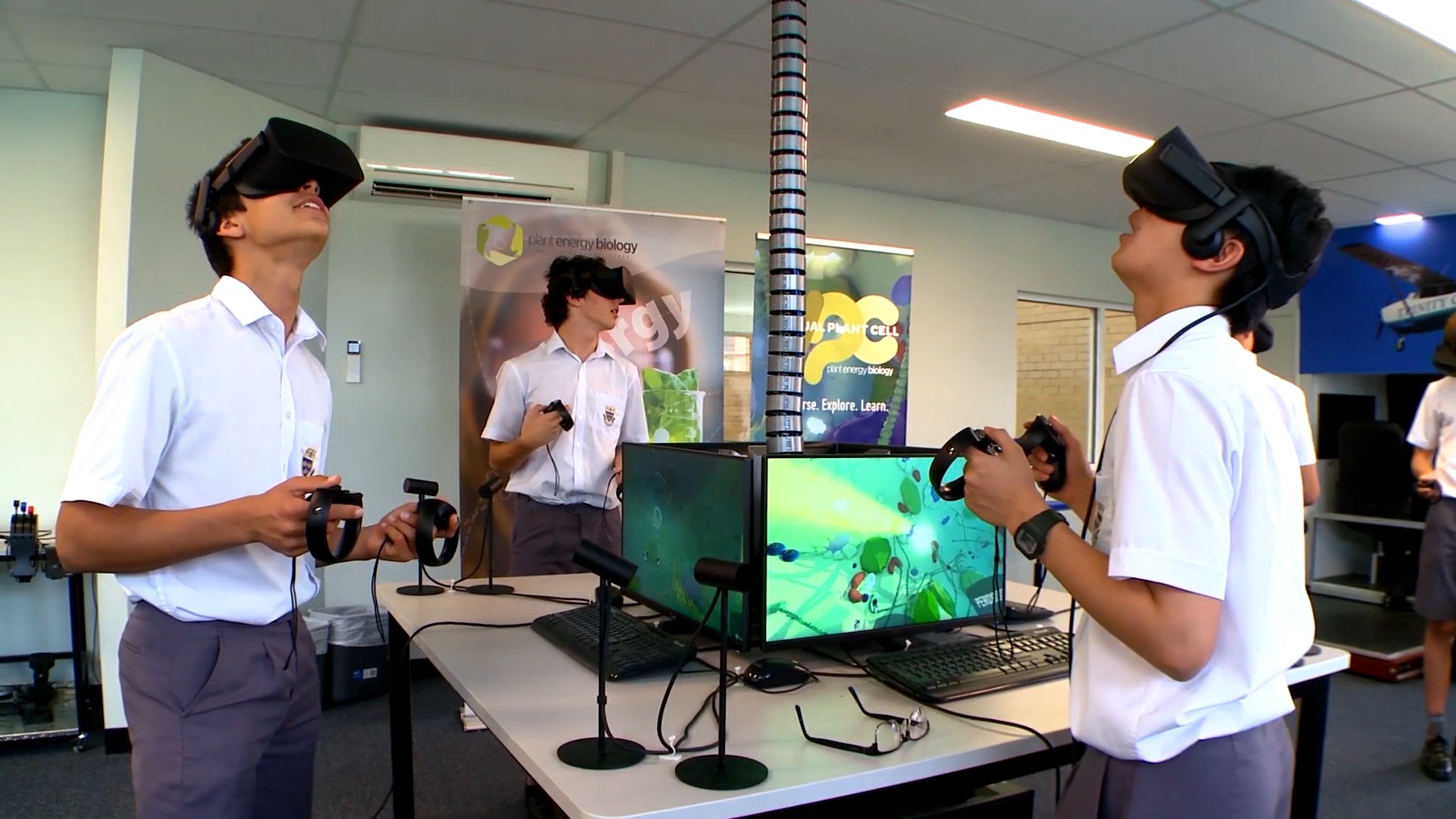 Two school students in VR headsets using Virtual Plant Cell: Classroom in a school lab, with the game on a monitor and a Virtual Plant Cell banner behind