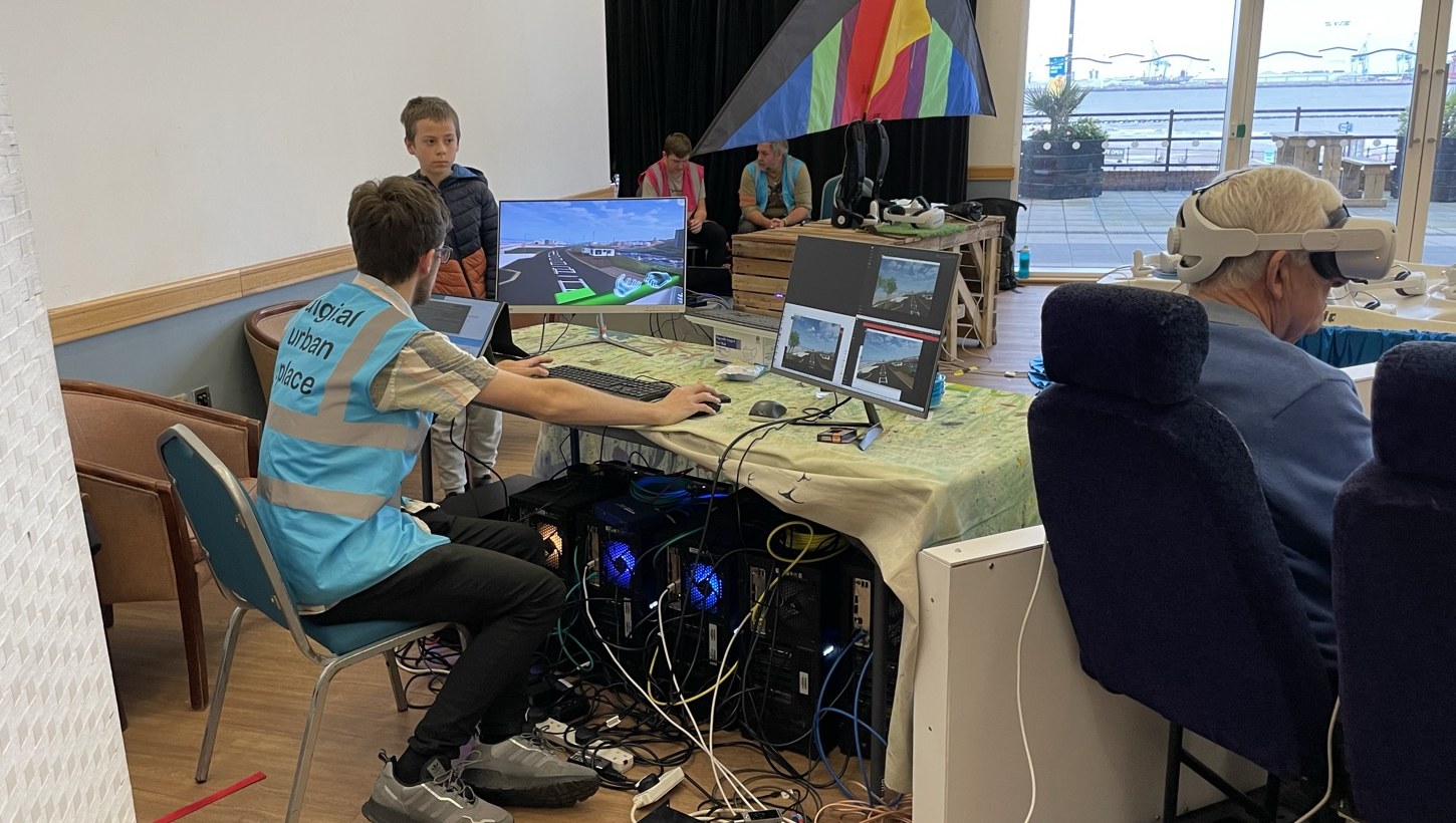 Behind the scenes at the New Brighton event: a crew member at the operators&rsquo; desk running the multi-user VR sessions, with screen views of the virtual promenade, while a visitor in a headset rides the bus rig