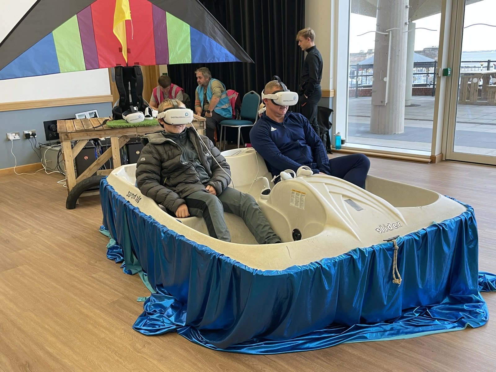 Two visitors in VR headsets pedalling a real pedalo, draped in blue fabric as water under a rainbow kite, at the Wirral consultation event