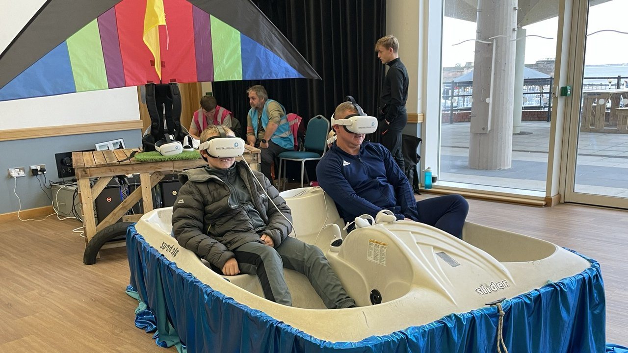 A boy and a man in VR headsets on a pedalo rig under a rainbow kite, at a VR public consultation in New Brighton